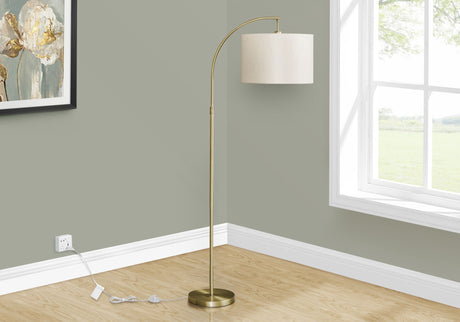The Contemporary Design Floor Lamp Lighting, featuring a curved brass-colored stand and a white cylindrical shade, sits by a large window on a wooden floor against a light green wall, with its power cord plugged into an outlet.