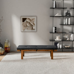 A minimalist living room features the Lucia black cushioned bench on a textured beige rug, accompanied by abstract wall art and a black metal shelf with white decor. Large jars with twigs sit on the floor to the left, creating a space dominated by neutral tones.