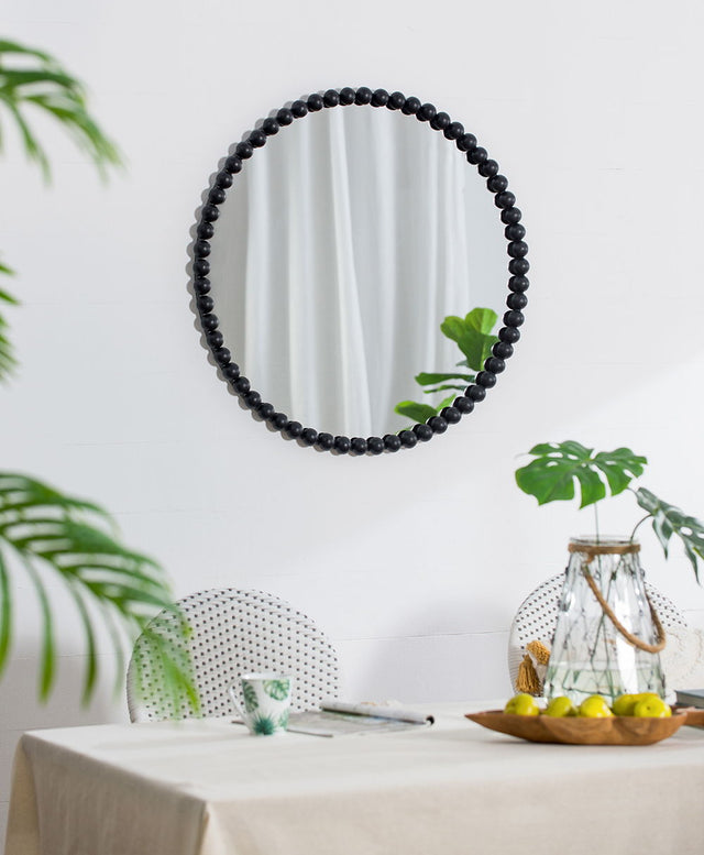 A Circle Mirror With Metal Beaded Frame - Black hangs on a white wall above a beige tablecloth-covered table with green printed chairs, lemons on a tray, a candle, and branches in a glass container. Reflected are green leaves and a curtain.