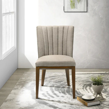 The Elmwood - Stylish Dining Chair with a beige upholstery and wooden legs sits on a patterned gray and white rug. Nearby, a small round table holds books, a plant, and cups. A framed leaf print adorns the textured wall as sunlight streams through a nearby window.