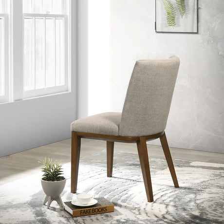 An Elmwood Stylish Dining Chair with a beige upholstered seat and wooden legs faces a bright window. Nearby, a small plant rests on "Fake Books." A light-colored patterned rug covers the floor, enhancing the minimalist decor.