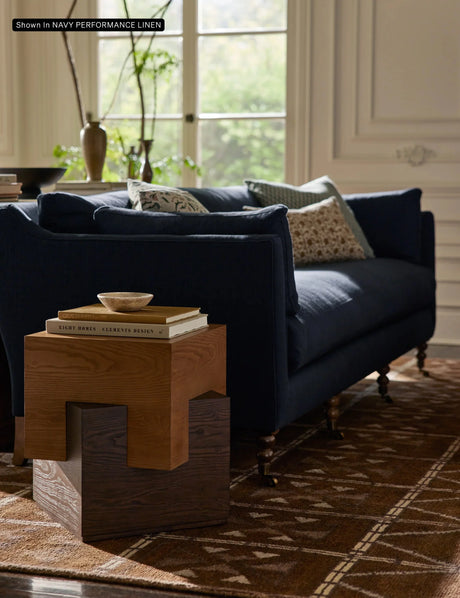 The living room features a cozy atmosphere with the Fabienne Sofa in navy, adorned with patterned cushions. A small wooden side table sits nearby, holding a few books and a decorative bowl. Natural light streams through a window showcasing lush green foliage outside, while a patterned rug adds warmth to the floor.