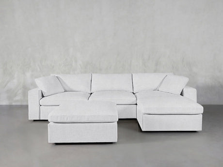 A contemporary, light gray 4-seat modular chaise sectional with an ottoman is elegantly arranged on a smooth gray floor, complemented by a textured gray wall. The sectional boasts plush cushions for added comfort.