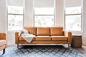 In a cozy living room, the Albany Sofa in vegan leather is adorned with a textured cream throw. Positioned against large windows, it allows natural light to flood the space. Nearby, a woven basket sits on a patterned blue rug.