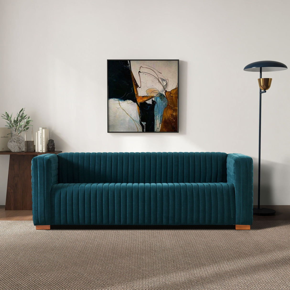 A modern living room features the Elara - Upholstered Sofa in teal against a white wall. An abstract painting with dark tones hangs above, a black floor lamp stands to the right, and a wooden console holding plants and books is on the left. The floor is carpeted.