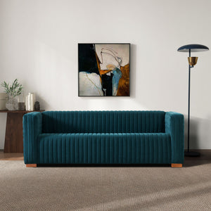 A modern living room features the Elara - Upholstered Sofa in teal against a white wall. An abstract painting with dark tones hangs above, a black floor lamp stands to the right, and a wooden console holding plants and books is on the left. The floor is carpeted.