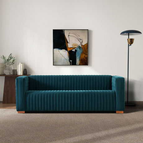A modern living room features the Elara - Upholstered Sofa in teal against a white wall. An abstract painting with dark tones hangs above, a black floor lamp stands to the right, and a wooden console holding plants and books is on the left. The floor is carpeted.