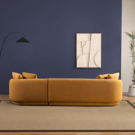 In a modern living room, the Chandler - Comfortable Sectional Sofa in mustard is adorned with cushions and sits against a dark blue wall. Above it hangs a minimalist abstract artwork. A sleek black floor lamp is on the left, while a potted plant sits to the right on a neutral rug.