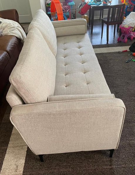 A light beige StyleWell Goodwin Mid-Century Modern Sofa with throw pillows and button tufted cushions is positioned next to a brown leather sofa. Children's toys and a small table with chairs can be seen in the cluttered background, all set against a carpeted floor.