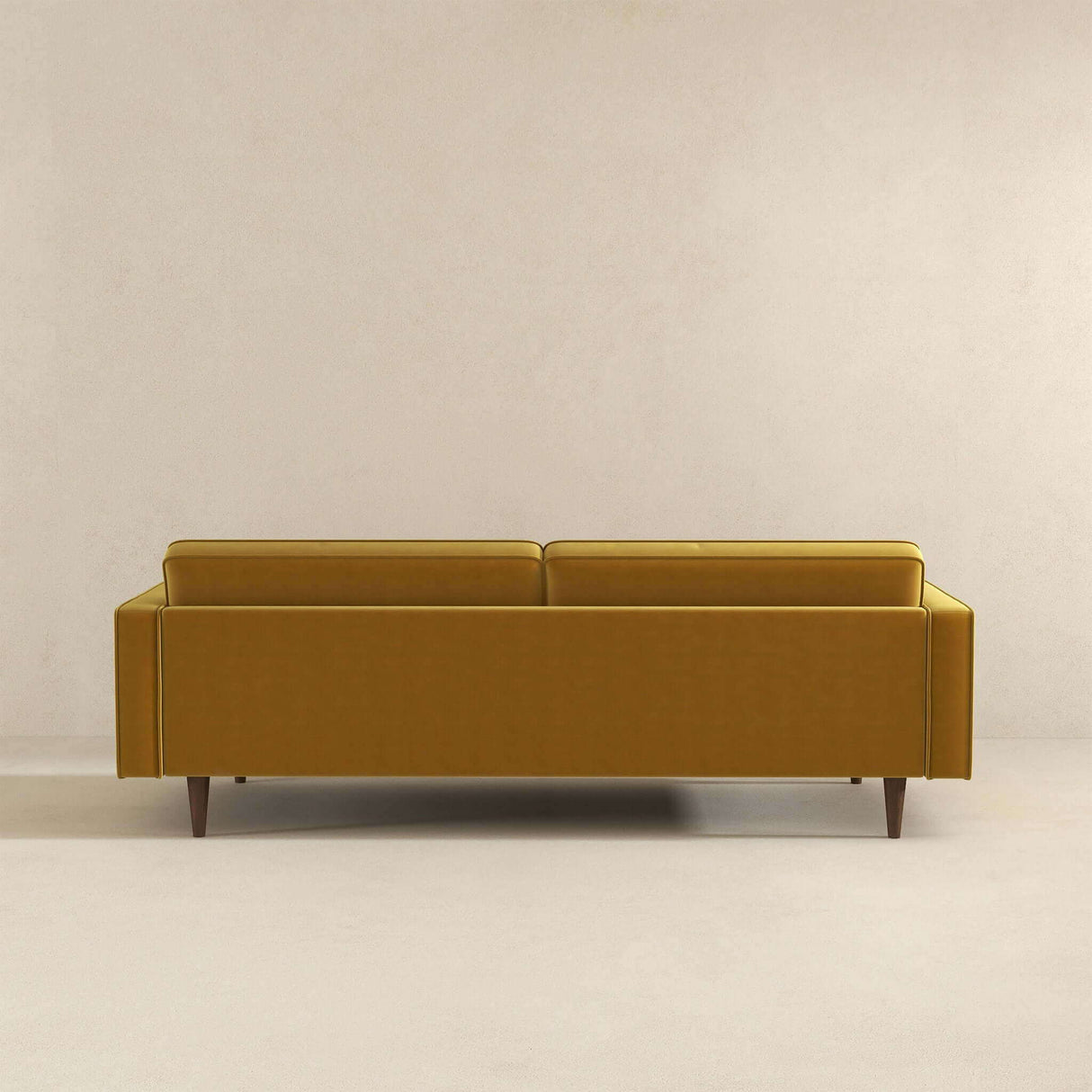 The Casey - Mid Century Modern Sofa in mustard yellow stands against a neutral beige wall, featuring a modern minimalistic design with clean lines and wooden legs. The image captures the back view of the sofa.