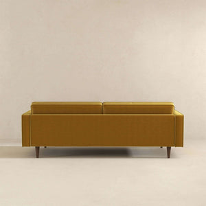 The Casey - Mid Century Modern Sofa in mustard yellow stands against a neutral beige wall, featuring a modern minimalistic design with clean lines and wooden legs. The image captures the back view of the sofa.