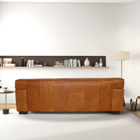 A cozy living room features a Morgan - Waxy Sofa in Cognac facing a white wall, with a narrow shelf behind displaying books and decor. A floor lamp stands to the right, while a small side table with books sits on the left. The floor is covered in cream carpet.