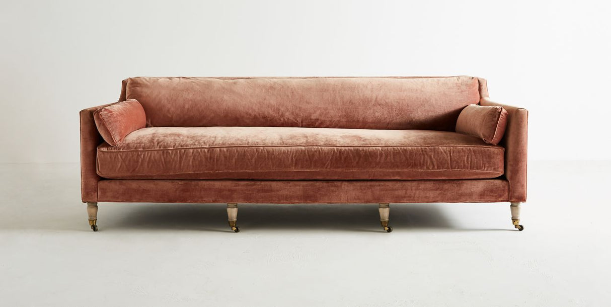The Leonelle Sofa is a contemporary, plush seating piece upholstered in luxurious, soft dusty rose velvet. It boasts a straight back design and includes two matching side pillows. The sofa stands on sleek wooden legs equipped with brass casters, all showcased against a minimalistic white backdrop.