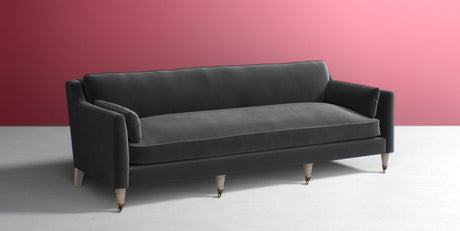 The Leonelle Sofa, featuring a minimalist design with clean lines and plush cushions, is placed against a stylish matte pink wall on a light gray floor. This modern gray sofa with wooden legs offers a comfortable and elegant seating option.