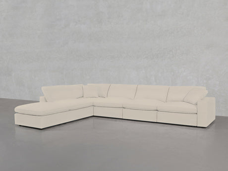 The 6-Seat Modular Corner Lounger Sectional features light beige upholstery, wide seat cushions, and pillow-style backrests, with a left-side chaise lounge. It sits on a gray concrete floor against a light gray textured wall.