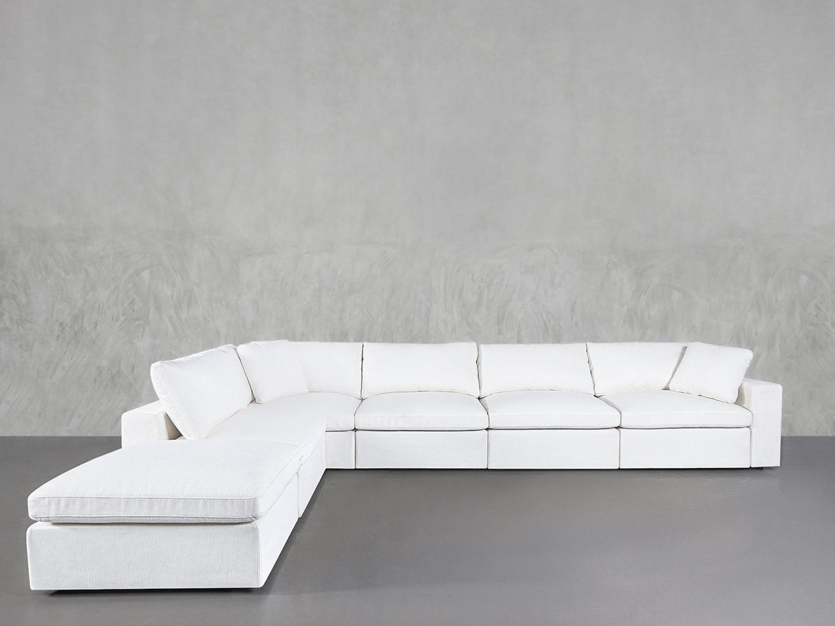The 6-Seat Modular Corner Lounger Sectional in white, with plush cushions, is arranged in an L-shape against a plain light gray textured wall and smooth gray floor.