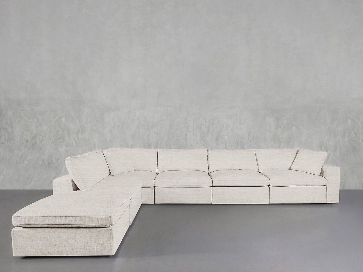 The 6-Seat Modular Corner Lounger Sectional in light beige, featuring clean lines and a chaise lounge on the left, sits against a textured gray wall and gray floor, showcasing a modern, minimalist design.
