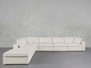 The 6-Seat Modular Corner Lounger Sectional in light beige, featuring clean lines and a chaise lounge on the left, sits against a textured gray wall and gray floor, showcasing a modern, minimalist design.