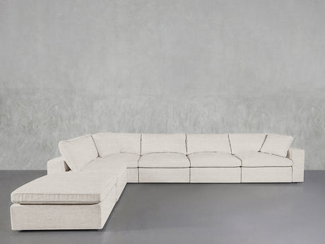 The 6-Seat Modular Corner Lounger Sectional in light beige, featuring clean lines and a chaise lounge on the left, sits against a textured gray wall and gray floor, showcasing a modern, minimalist design.