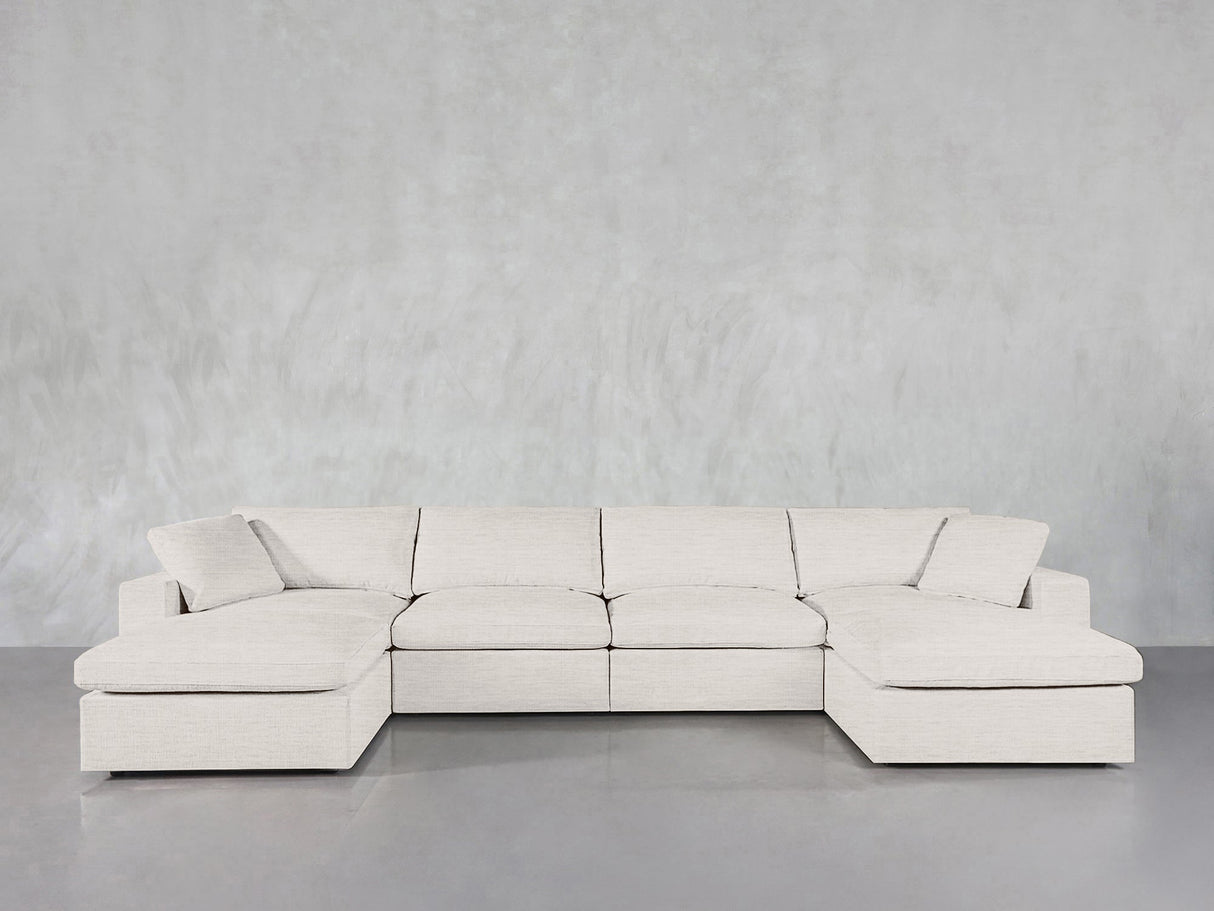 A 6-Seat Modular Double Chaise Sectional in light beige with clean lines and plush cushions sits against a textured gray wall on a smooth gray floor.