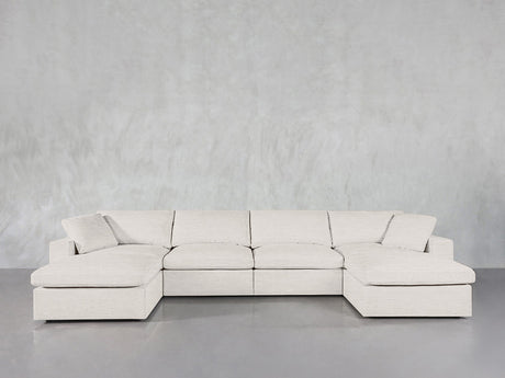 A 6-Seat Modular Double Chaise Sectional in light beige with clean lines and plush cushions sits against a textured gray wall on a smooth gray floor.