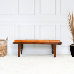 The Maddox bench in rustic tan sits against a white paneled wall. A woven basket and a black pot with tall dried grass adorn the light carpeted floor beside it.