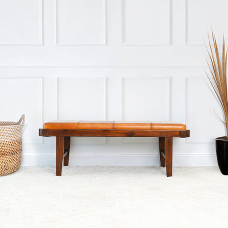 The Maddox bench in rustic tan sits against a white paneled wall. A woven basket and a black pot with tall dried grass adorn the light carpeted floor beside it.