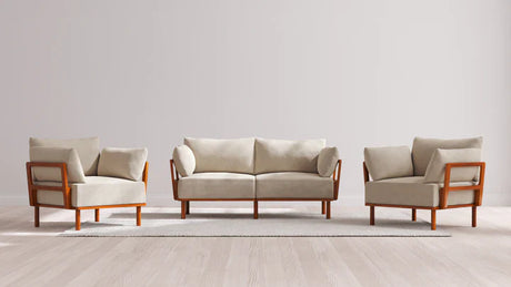 A minimalist living room set featuring the Indoor Jack 2-Seater Sofa and Armchair Set in Balsa, complete with beige cushions and wooden frames, arranged on a light-colored area rug over a wood floor, against a plain gray background.
