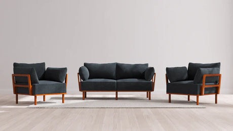 The Indoor Jack 2-Seater Sofa + Armchair Set in Balsa features a sleek and modern design with a dark gray finish. It includes a sofa and two matching armchairs with elegant wooden frames, all arranged on a light-colored rug atop a wooden floor, set against a plain, light gray wall.