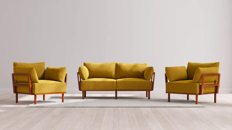 A stylish living room arrangement featuring the Indoor Jack 2-Seater Sofa + Armchair Set in Balsa, showcasing a mustard yellow sofa accompanied by two matching armchairs. They rest elegantly on a light beige rug over a light wood floor, with wooden frames accentuating the furniture against a plain wall backdrop.
