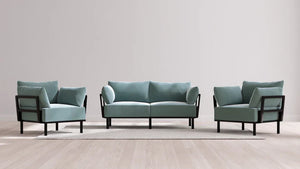 A contemporary living room setup showcasing the Indoor Jack 2-Seater Sofa + Armchair Set in Balsa, featuring teal cushions and black frames. The set is displayed on a light-colored rug placed over a wooden floor, against a plain white wall backdrop.