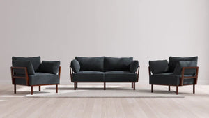 The Indoor Jack 2-Seater Sofa and Armchair Set in Balsa showcases a sleek design with a dark gray sofa and two matching armchairs, all featuring elegant wooden frames. They are placed on a light-colored rug on top of a wooden floor, complementing the simplicity of a plain white wall.