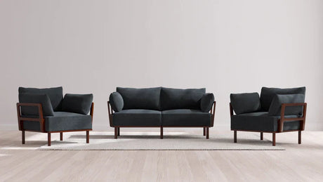 The Indoor Jack 2-Seater Sofa and Armchair Set in Balsa showcases a sleek design with a dark gray sofa and two matching armchairs, all featuring elegant wooden frames. They are placed on a light-colored rug on top of a wooden floor, complementing the simplicity of a plain white wall.
