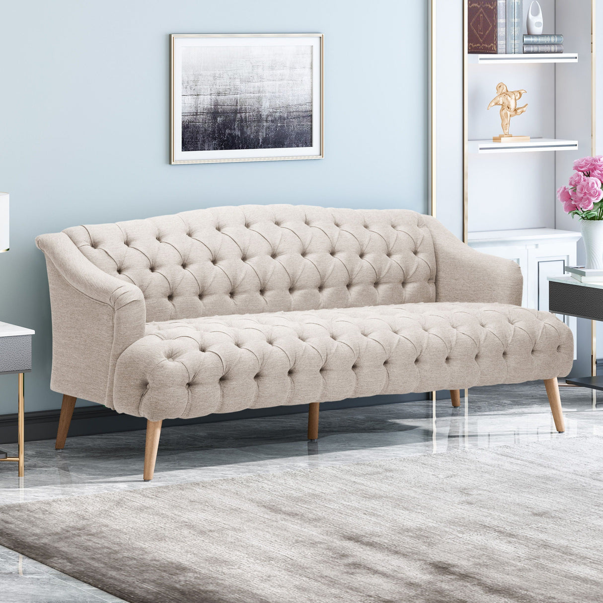 The Comfy 3 Seat Sofa with wooden legs sits in a modern living room, complemented by a framed abstract artwork on the wall. Decorative shelves on the right hold books, a small sculpture, and a vase of pink flowers, while a textured rug covers the floor.