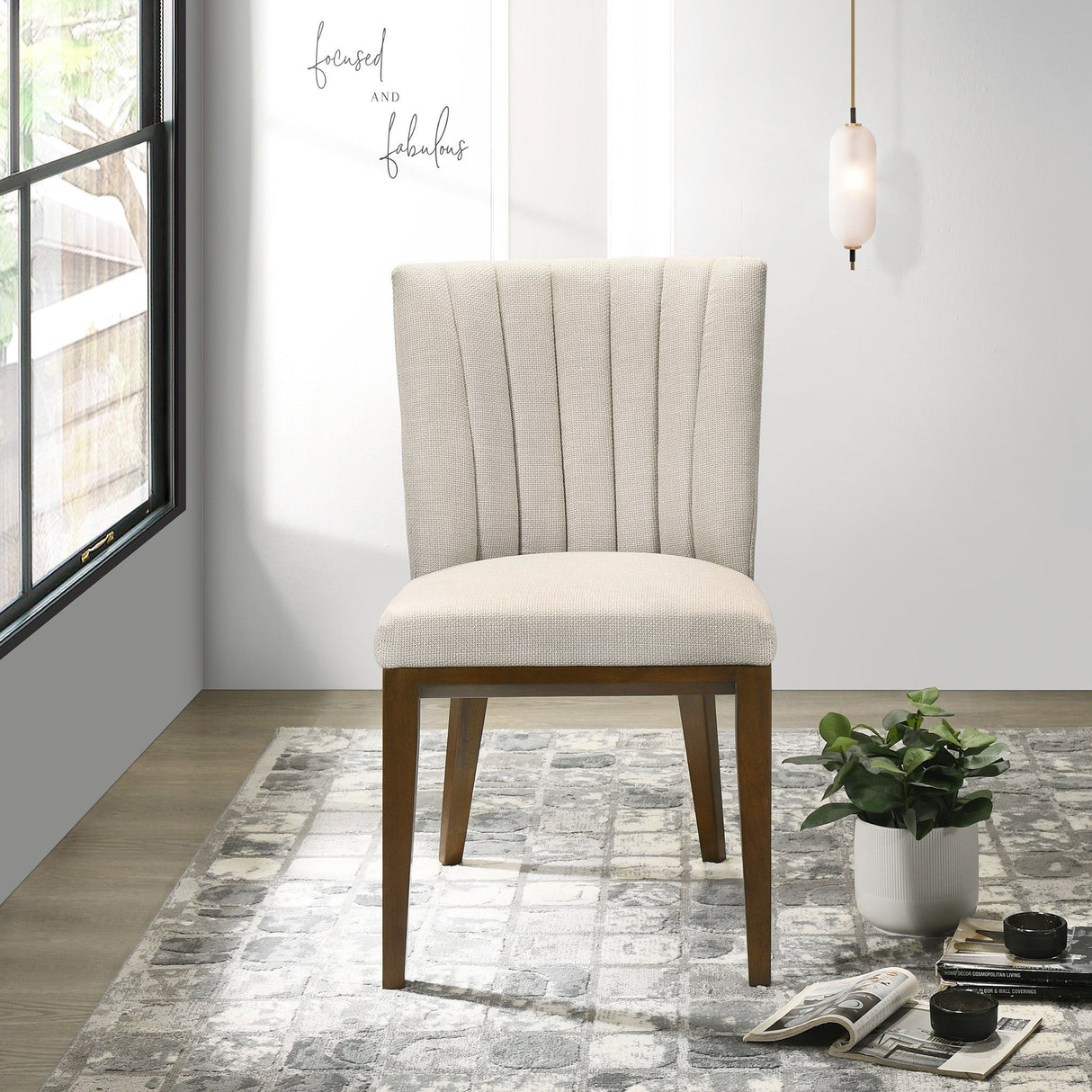 A beige upholstered Elmwood dining chair with a wooden frame sits on a patterned gray rug. Nearby, an open magazine and a small plant are on the floor. The minimalist space is adorned with wall art saying "focused and fabulous," along with a window and pendant light.