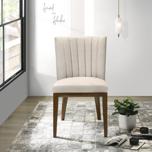 A beige upholstered Elmwood dining chair with a wooden frame sits on a patterned gray rug. Nearby, an open magazine and a small plant are on the floor. The minimalist space is adorned with wall art saying "focused and fabulous," along with a window and pendant light.