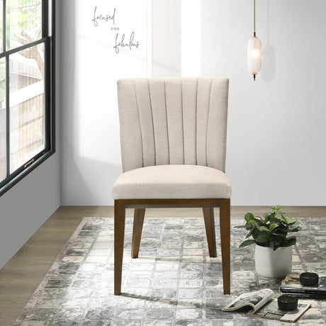 A beige upholstered Elmwood dining chair with a wooden frame sits on a patterned gray rug. Nearby, an open magazine and a small plant are on the floor. The minimalist space is adorned with wall art saying "focused and fabulous," along with a window and pendant light.