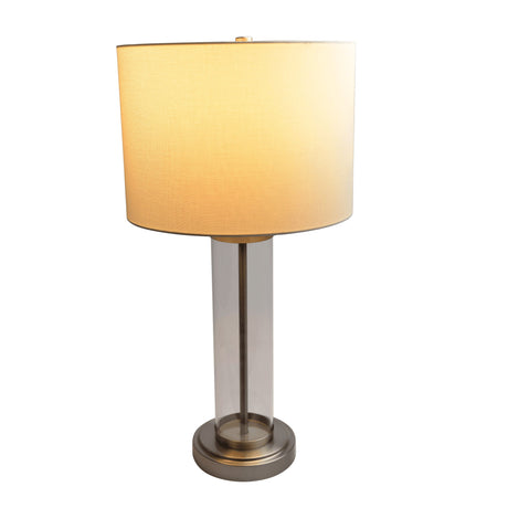The Luna table lamp features a clear and silver cylindrical glass and metal base, topped with a beige round fabric lampshade. It emits warm light and stands on a circular metal base.