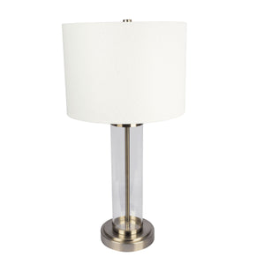 The Luna table lamp features a cylindrical clear glass base with a brushed metal bottom, white drum shade, and central metal rod topped by a metallic finial.