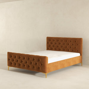 The Bailey - Elegant Design Bed features a tufted, caramel-toned headboard and footboard, offering stylish elegance in a minimalist, light-colored room. It boasts an upholstered frame supported by slender gold legs.