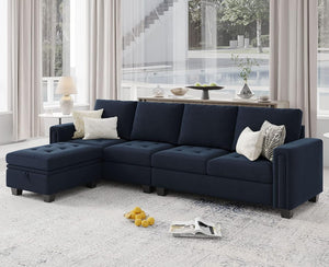 A modern living room showcases a spacious Belffin Reversible Sectional Convertible Storage sofa in deep blue, perfectly contrasted by a light gray carpet. The sofa is adorned with various white and patterned throw pillows. In the background, a console table with decorative items is positioned in front of tall windows draped with sheer curtains.