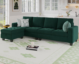 A modern living room features the Belffin Reversible Sectional Convertible Storage in dark green, adorned with striped and patterned pillows. A matching ottoman rests nearby on a light rug showcasing green geometric designs. The room is accentuated by large windows, light-colored walls, and offers a glimpse of the kitchen area.