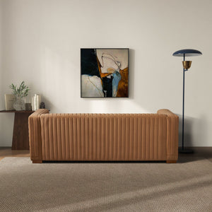 A modern living room features the Elara - Upholstered Sofa, a tall black and gold floor lamp, and a side table adorned with books and a plant. An abstract painting decorates the light-colored wall, while a neutral-toned rug covers the floor.