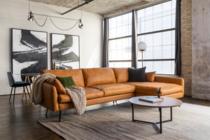 A modern living room featuring the Park Sectional Sofa in tan leather, enhanced with textured pillows and a knit throw. The space is complemented by a round wooden coffee table and abstract black and white paintings, with large windows that provide natural light.
