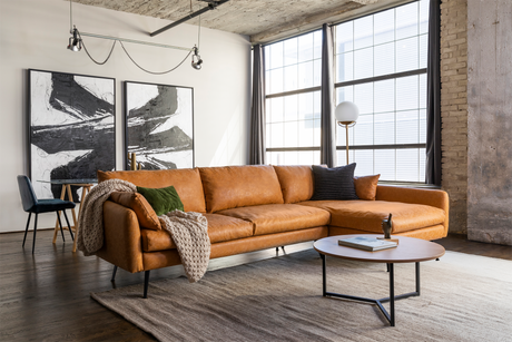 A modern living room featuring the Park Sectional Sofa in tan leather, enhanced with textured pillows and a knit throw. The space is complemented by a round wooden coffee table and abstract black and white paintings, with large windows that provide natural light.