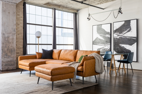 A stylish living room featuring the spacious Park Sectional Sofa in tan leather, accompanied by a matching ottoman on a gray rug. Minimalist black and white artwork adorns the walls, while a modern floor lamp and a compact dining table with chairs are situated nearby. Large windows provide abundant natural light.