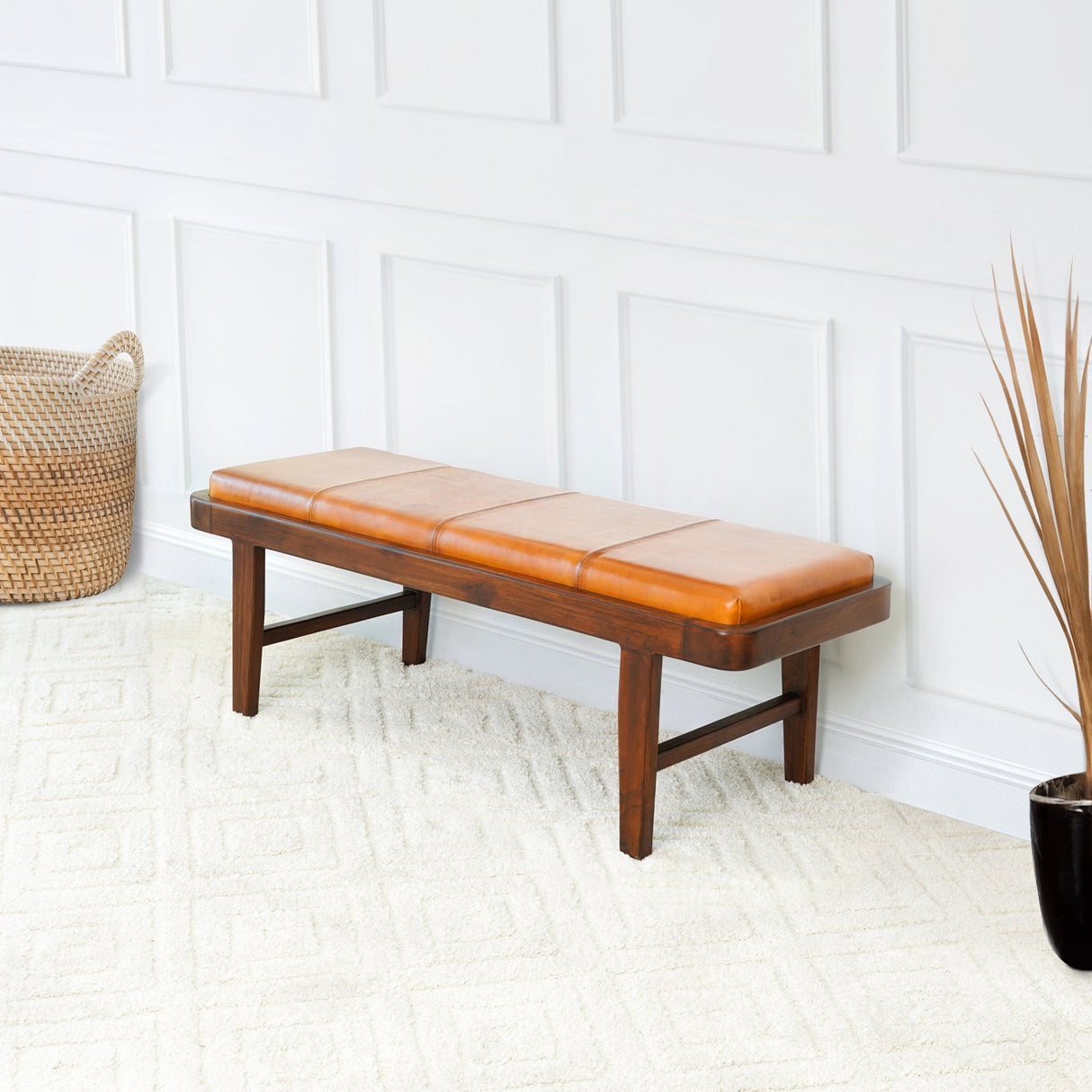 The Maddox bench in rustic tan features a cushioned seat, resting on a textured cream carpet. Nearby, a woven basket accents the corner while tall dried grasses complement the white paneled wall.
