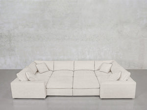 The 8-Seat Modular Closed Pit, a spacious light beige sectional with four matching cushions, is arranged against a plain light grey concrete wall on a smooth grey floor.