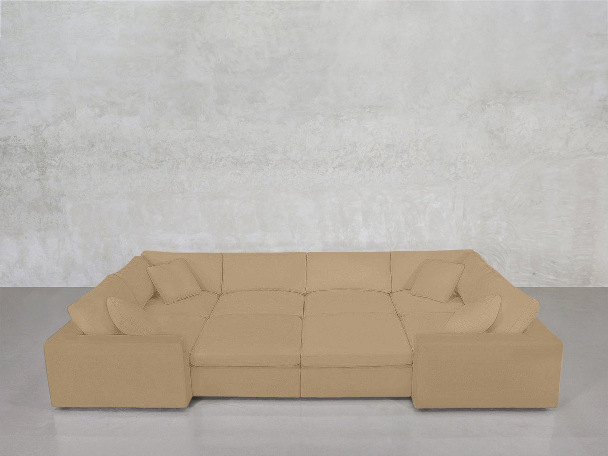 An 8-Seat Modular Closed Pit sectional in beige, featuring wide armrests, a modern design, and four matching throw pillows, sits against a light gray textured wall on a polished concrete floor.