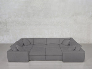The 8-Seat Modular Closed Pit is a large, modern gray sectional with wide armrests, four matching cushions, and a minimalist design, set against a light gray textured wall and smooth gray floor.